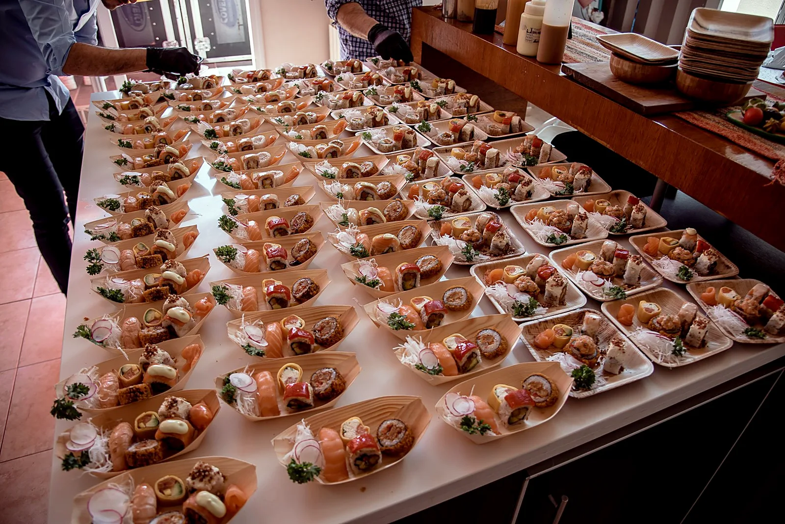 Mesa larga con decenas de barquitos de madera con rolls, niguiris y sashimi listos para servir en un evento, mientras el equipo termina de armarlos