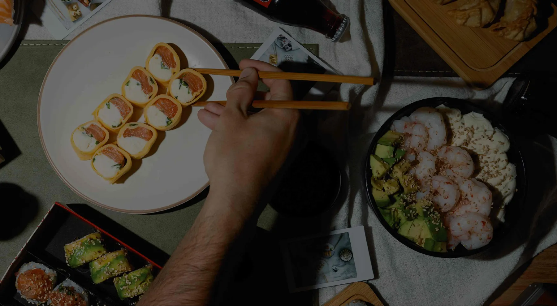 Mesa de sushi vista desde arriba: rolls de salmón en un plato, una salad de langostinos con palta y una mano que toma una pieza con palitos