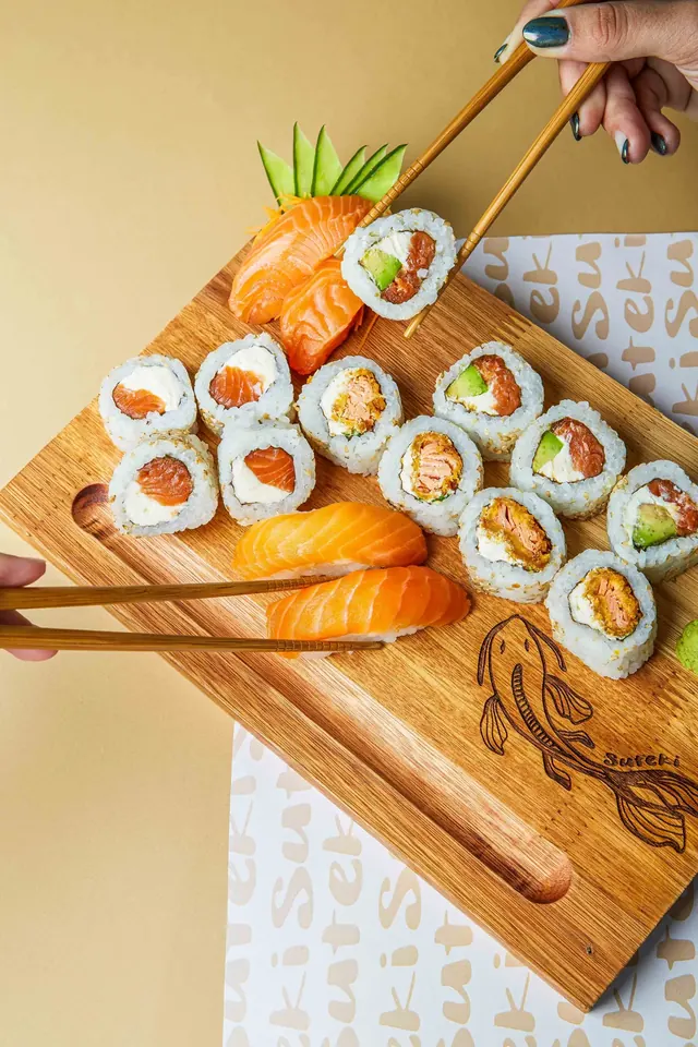 Tabla de madera de Suteki con rolls de salmón, palta y philadelphia, rolls crispy, niguiris y sashimi de salmón, y palitos tomando una pieza